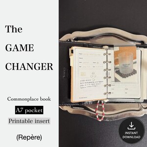 May include: A black A7 pocket binder with a six-ring metal mechanism holding white paper inserts. The pages feature printed project tracking fields and a small photograph of a glass mug. A beaded charm with pink and white beads hangs from the rings.