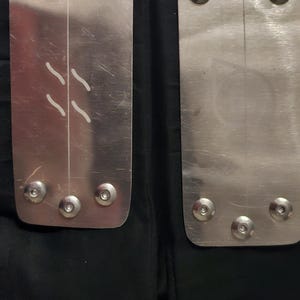 May include: Two silver metal headbands with a rectangular shape and rounded corners. Each headband has six silver screws and a white line down the center. One headband has two white diagonal lines.
