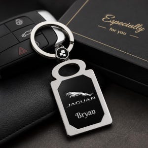 May include: A silver and black Jaguar keyring featuring the Jaguar logo and the name "Bryan". The keyring is attached to a key ring and is next to a car key and a black gift box with the words "Especially for you".