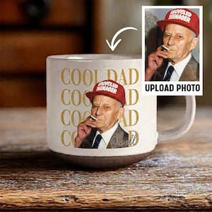 May include: A white ceramic mug with a brown base features a photo of an older man wearing a red cap that says "recycled teenager." The mug also has the words "COOL DAD" in gold lettering. The mug has a handle.