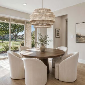 May include: A dining room set featuring a round wooden table and six cream-colored upholstered chairs. A large, layered woven pendant light hangs above the table. A vase with greenery sits on the table. Large windows offer a view of the outdoors.