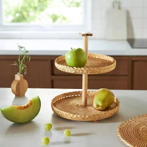 May include: A two-tiered woven rattan serving tray with a wooden center post. The top tier holds a green apple, and the bottom tier holds a pear. The tray is on a white countertop with a vase and a slice of melon.