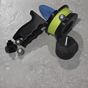 May include: A black and blue fishing reel with a yellow line, a black handle, and silver weights. The reel is designed for fishing and is made of plastic. The reel is on a gray surface.