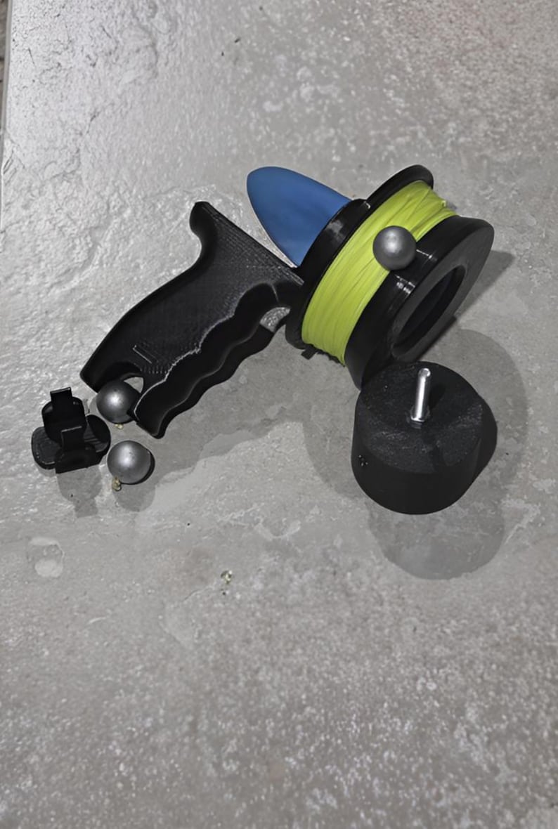 May include: A black and blue fishing reel with a yellow line, a black handle, and silver weights. The reel is designed for fishing and is made of plastic. The reel is on a gray surface.