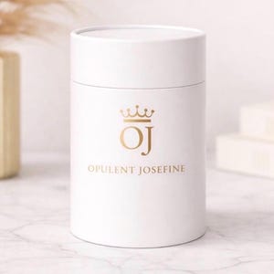 Könnte beinhalten: Ein weißer zylindrischer Behälter mit passendem Deckel. Der Behälter hat ein goldenes Logo mit den Buchstaben "OJ", gekrönt von einer Krone, und die Worte "OPULENT JOSEFINE" in goldener Schrift. Der Behälter steht auf einer Marmoroberfläche.