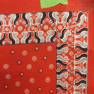 May include: Red fabric with a white and black patterned border. The border features a repeating design of stylized figures. The fabric has a scattered pattern of small white and black circles.