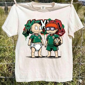K&ouml;nnte beinhalten: Ein cremefarbenes Kurzarm-T-Shirt mit einer Grafik von zwei Zeichentrickfiguren in gr&uuml;nen Fu&szlig;balltrikots. Eine Figur tr&auml;gt eine Windel, die andere h&auml;lt einen Fu&szlig;ball. Dahinter steht das Wort MEXICO in Graffiti-Schrift mit roten, wei&szlig;en und gr&uuml;nen Akzenten.