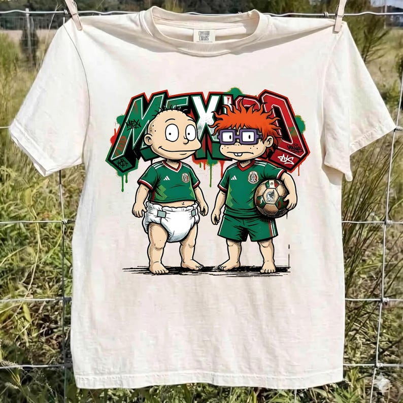 K&ouml;nnte beinhalten: Ein cremefarbenes Kurzarm-T-Shirt mit einer Grafik von zwei Zeichentrickfiguren in gr&uuml;nen Fu&szlig;balltrikots. Eine Figur tr&auml;gt eine Windel, die andere h&auml;lt einen Fu&szlig;ball. Dahinter steht das Wort MEXICO in Graffiti-Schrift mit roten, wei&szlig;en und gr&uuml;nen Akzenten.