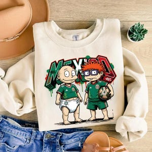 K&ouml;nnte beinhalten: Ein cremefarbenes Sweatshirt mit Rundhalsausschnitt und einer Grafik von zwei Zeichentrickfiguren in gr&uuml;nen Fu&szlig;balltrikots. Eine Figur tr&auml;gt eine Windel, die andere h&auml;lt einen Fu&szlig;ball. Das Wort MEXICO ist in gro&szlig;en, stilisierten Graffiti-Buchstaben in Rot, Wei&szlig; und Gr&uuml;n dahinter gedruckt.