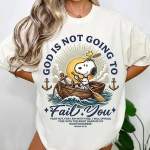 May include: Cream-coloured t-shirt with Snoopy in a boat, and the text "GOD IS NOT GOING TO FAIL YOU." The design includes anchors, waves, and a sun. The quote "FEAR NOT, FOR I AM WITH THEE, I WILL UPHOLD THEE WITH THE RIGHT HAND OF MY RIGHTEOUSNESS. ISAIAH 41:10" is also included.
