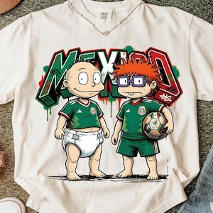 K&ouml;nnte beinhalten: Ein cremefarbenes T-Shirt mit einer Grafik von zwei Zeichentrickfiguren in gr&uuml;nen Mexiko-Fu&szlig;balltrikots. Eine Figur tr&auml;gt eine Windel, die andere h&auml;lt einen Fu&szlig;ball. Dahinter steht das Wort MEXICO in gro&szlig;en Buchstaben im Graffiti-Stil.