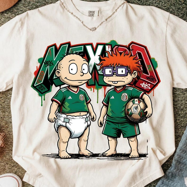 K&ouml;nnte beinhalten: Ein cremefarbenes T-Shirt mit einer Grafik von zwei Zeichentrickfiguren in gr&uuml;nen Mexiko-Fu&szlig;balltrikots. Eine Figur tr&auml;gt eine Windel, die andere h&auml;lt einen Fu&szlig;ball. Dahinter steht das Wort MEXICO in gro&szlig;en Buchstaben im Graffiti-Stil.