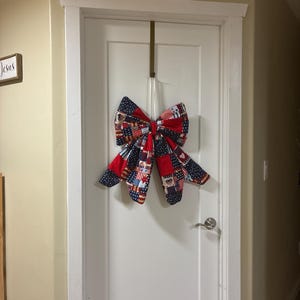 Op de afbeelding: Een grote decoratieve strik van stof met een patchworkpatroon in rood, wit en blauw, voorzien van sterren- en strepenmotieven. De strik hangt aan een dun lint aan een metalen haak boven op een witte binnendeur.
