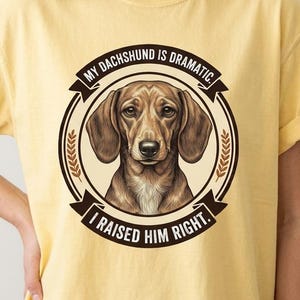 Pode incluir: Uma t-shirt de manga curta em amarelo pálido com um gráfico circular que apresenta uma ilustração de um cão dachshund castanho. O texto diz: "MY DACHSHUND IS DRAMATIC. I RAISED HIM RIGHT." O design é emoldurado por faixas castanhas escuras.