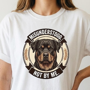 May include: A white short-sleeved t-shirt featuring a circular graphic on the chest. The graphic displays a Rottweiler dog portrait centered within a tan circle, surrounded by a dark brown border with the text MISUNDERSTOOD NOT BY ME.