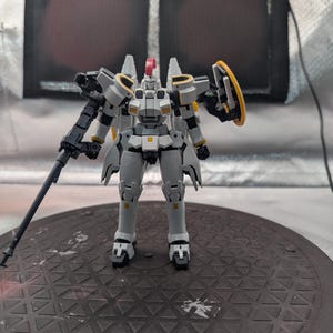 以下が含まれることがあります： 黄色と赤のアクセントが付いた、詳細なグレーと白のモデルロボット。 ロボットは片方の腕に大きな銃、もう一方の腕にシールドを持っています。 濃い灰色のテクスチャのある表面に立っています。