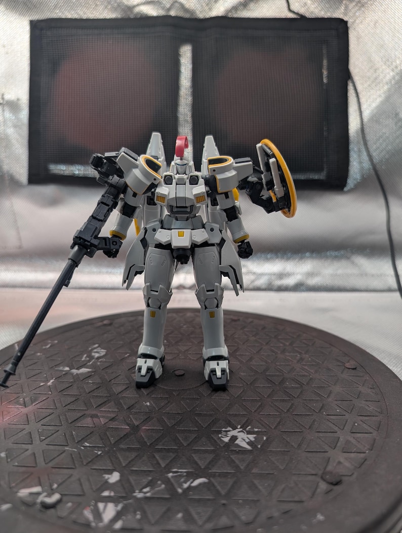 以下が含まれることがあります： 黄色と赤のアクセントが付いた、詳細なグレーと白のモデルロボット。 ロボットは片方の腕に大きな銃、もう一方の腕にシールドを持っています。 濃い灰色のテクスチャのある表面に立っています。