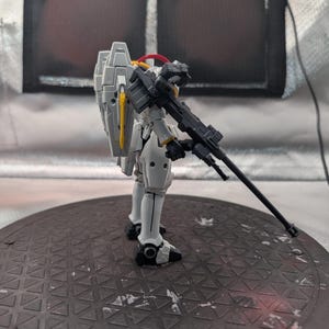 以下が含まれることがあります： 黒いライフルを持った白とグレーのモデルロボットフィギュア。フィギュアは黄色と赤のアクセントがあり、濃い灰色の円形のベースに立っています。背景は、濃い灰色の正方形の明るい灰色です。