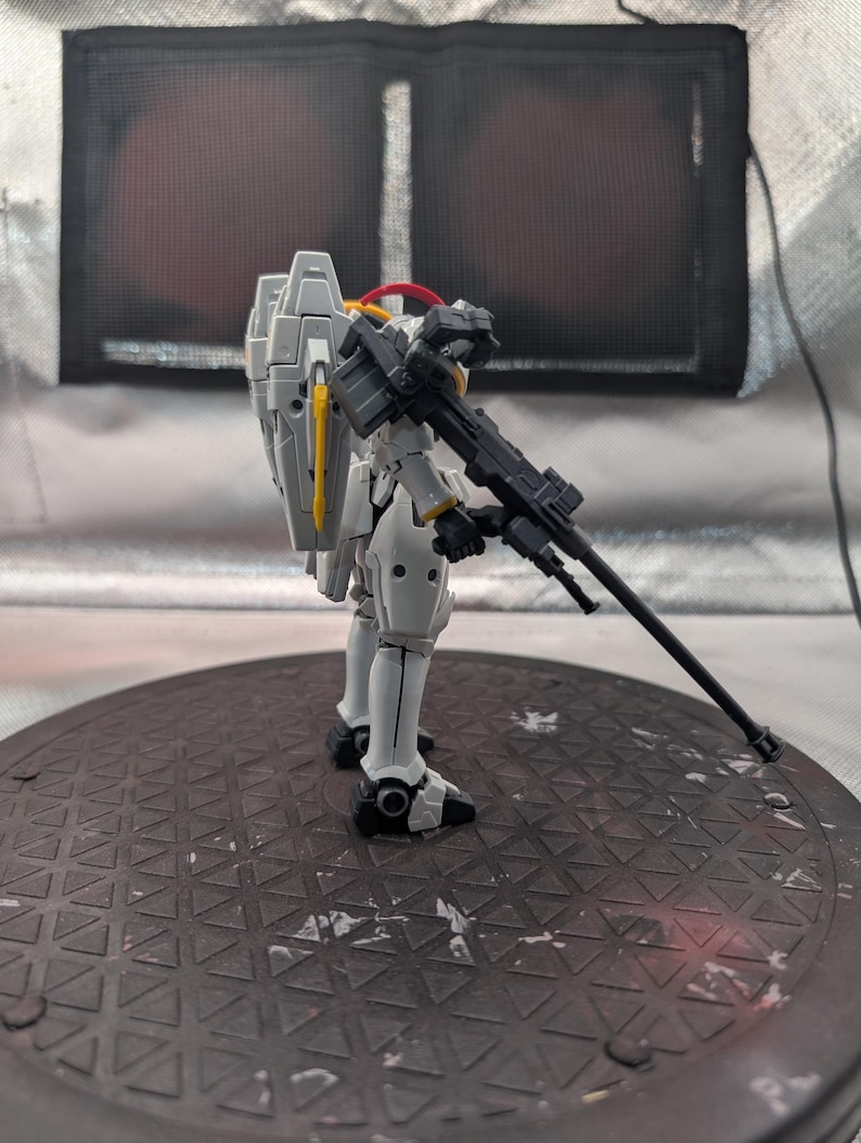 以下が含まれることがあります： 黒いライフルを持った白とグレーのモデルロボットフィギュア。フィギュアは黄色と赤のアクセントがあり、濃い灰色の円形のベースに立っています。背景は、濃い灰色の正方形の明るい灰色です。