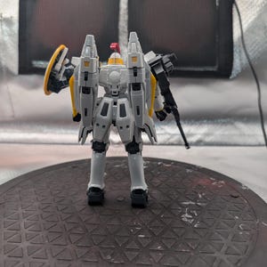 以下が含まれることがあります： 黒いライフルと黄色の盾を持った、黄色と赤のアクセントが付いたグレーと白のモデルロボット。ロボットは、テクスチャ加工された濃い灰色の表面に立っています。