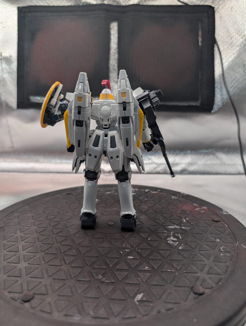 以下が含まれることがあります： 黒いライフルと黄色の盾を持った、黄色と赤のアクセントが付いたグレーと白のモデルロボット。ロボットは、テクスチャ加工された濃い灰色の表面に立っています。