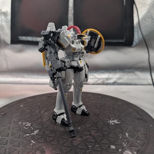 以下が含まれることがあります： 白とグレーのロボットアクションフィギュア。黄色と赤のアクセントが付いています。フィギュアはグレーの武器と黄色の盾を持っています。ロボットは濃い灰色の表面に立っています。