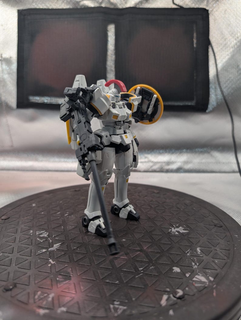 以下が含まれることがあります： 白とグレーのロボットアクションフィギュア。黄色と赤のアクセントが付いています。フィギュアはグレーの武器と黄色の盾を持っています。ロボットは濃い灰色の表面に立っています。