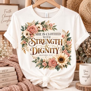 May include: A white short-sleeved t-shirt featuring a floral wreath graphic surrounding the text "SHE IS CLOTHED WITH STRENGTH & DIGNITY PROVERBS 31:25". The design includes pink roses, a large yellow sunflower, and green leaves on a crew neck shirt.