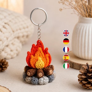 May include: A crochet campfire keychain featuring a silver metal ring and chain. The base consists of gray crocheted stones, brown crocheted logs, and a central flame made of layered yellow, orange, and red yarn. The design is compact and textured.