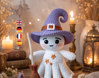 Könnte beinhalten: Eine weiße gehäkelte Geisterpuppe mit einem violetten spitzen Hut, einem braunen Band und einer weißen Schnalle. Der Geist hat schwarze Augen, rosa Wangen und kleine orangefarbene Sternstickereien auf der Brust. Er hält einen kleinen braunen Besen.