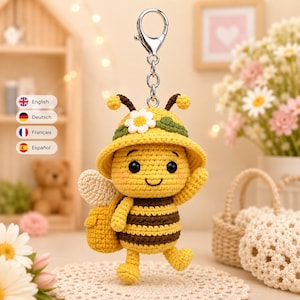 Puede incluir: Un llavero de abeja tejido a ganchillo con cuerpo a rayas amarillas y marrones, sombrero amarillo con una flor blanca y pequeñas alas blancas. La abeja tiene ojos negros, una sonrisa bordada y mejillas rosadas. Unido a un mosquetón de metal.