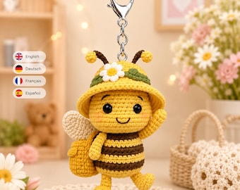 Könnte beinhalten: Ein gehäkelter Bienen-Schlüsselanhänger mit gelb-braun gestreiftem Körper, einem gelben Hut mit weißer Blume und kleinen weißen Flügeln. Die Biene hat schwarze Augen, ein aufgesticktes Lächeln und rosa Wangen. Befestigt an einem silbernen Karabiner.