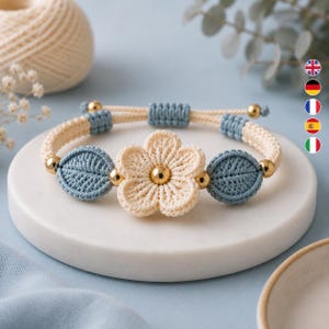 May include: A macrame bracelet featuring a central cream-colored flower with a gold bead center, flanked by two blue leaf-shaped macrame elements. The band is constructed from cream and blue cord with gold accent beads and an adjustable closure.