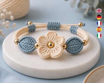 May include: A macrame bracelet featuring a central cream-coloured flower with a gold bead centre, flanked by two blue leaf-shaped macrame elements. The band is constructed from cream and blue cord with gold accent beads and an adjustable closure.