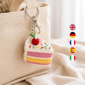 May include: A crochet keyring shaped like a slice of cake with a red cherry and a white candle on top. The cake features layers of cream, pink, and yellow yarn, decorated with colourful sprinkles. It is attached to a silver metal keyring and clasp.
