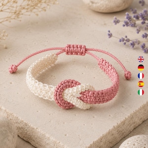 May include: A woven cord bracelet featuring a central knot design. The bracelet is constructed from alternating sections of cream and dusty rose colored thread. It includes an adjustable sliding closure mechanism with two small knotted ends.
