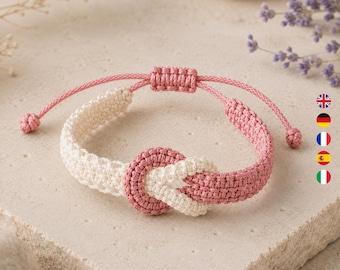 Könnte beinhalten: Ein geflochtenes Kordelarmband mit einem zentralen Knotendesign. Das Armband besteht aus abwechselnden Abschnitten aus cremefarbenem und altrosa farbenem Garn. Es verfügt über einen verstellbaren Schiebeverschluss mit zwei kleinen geknoteten Enden.