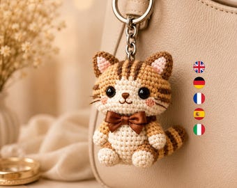 Könnte beinhalten: Ein gehäkelter Katzen-Schlüsselanhänger mit einem Körper aus braunem und cremefarbenem Garn, schwarzen Kunststoffaugen, rosa gestickten Wangen und einer dunkelbraunen Satinschleife. Die Katze hängt an einem silberfarbenen Ring an einer beigen Tasche.