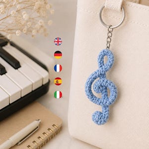 May include: A light blue crochet treble clef keychain attached to a cream-colored canvas tote bag. The keychain features a silver-toned metal ring and chain, showcasing a textured, yarn-based musical symbol design in a soft, pastel blue shade.