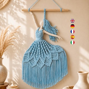 Könnte beinhalten: Ein Makramee-Wandbehang, der eine Figur in einem hellblauen Kleid mit komplexen Knotenmustern und langen Fransen darstellt. Die Figur hat cremefarbene Arme und einen Kopf und ist an einem horizontalen Holzstab zur Aufhängung befestigt.