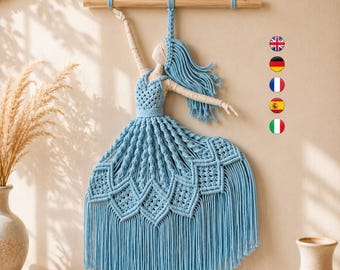 Könnte beinhalten: Ein Makramee-Wandbehang, der eine Figur in einem hellblauen Kleid mit komplexen Knotenmustern und langen Fransen darstellt. Die Figur hat cremefarbene Arme und einen Kopf und ist an einem horizontalen Holzstab zur Aufhängung befestigt.