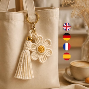 May include: A cream-colored canvas tote bag featuring a macrame keychain attachment. The keychain consists of a gold-toned metal ring, a long cream-colored cord tassel with a gold bead, and a cream-colored macrame flower with a yellow center.