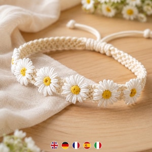 May include: A cream-colored macrame bracelet featuring a series of five white daisy flowers with yellow centers. The bracelet is constructed with a woven cord pattern and includes an adjustable sliding knot closure with two dangling cord ends.
