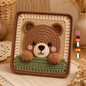 May include: A square crochet wall hanging featuring a brown bear face with black eyes, a dark brown nose, and pink cheeks. The bear is peeking over a green textured border. The entire piece is framed by a cream-coloured square border with a dark brown edge.