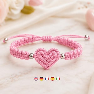 May include: A pink macrame bracelet featuring a central woven heart design. The cord is crafted from pink thread and includes two small silver-toned metallic beads flanking the heart, with adjustable sliding knots and silver-toned end beads.