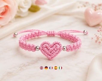 Könnte beinhalten: Ein rosafarbenes Makramee-Armband mit einem zentralen, geflochtenen Herzmotiv. Das Band besteht aus rosa Garn und enthält zwei kleine silberfarbene Metallperlen neben dem Herz sowie verstellbare Schiebeknoten und silberfarbene Endperlen.