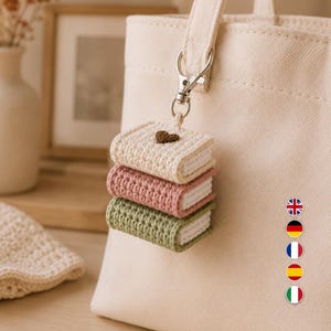 Puede incluir: Un bolso de tela color crema con un accesorio de llavero de ganchillo. El llavero consta de tres libros en miniatura apilados en color crema, rosa empolvado y verde salvia, con un peque&ntilde;o coraz&oacute;n marr&oacute;n en el libro superior, unido por un cierre plateado.