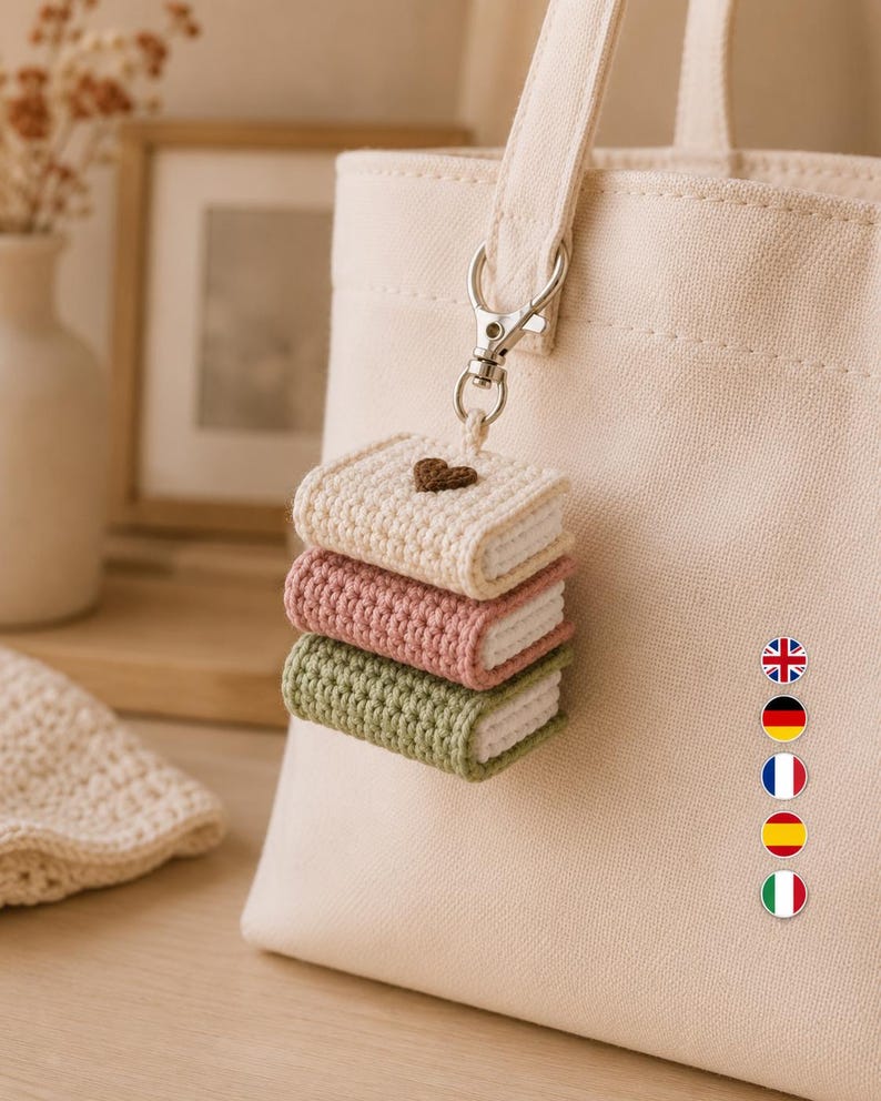 Puede incluir: Un bolso de tela color crema con un accesorio de llavero de ganchillo. El llavero consta de tres libros en miniatura apilados en color crema, rosa empolvado y verde salvia, con un peque&ntilde;o coraz&oacute;n marr&oacute;n en el libro superior, unido por un cierre plateado.
