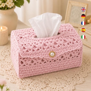 Puede incluir: Una funda rectangular de ganchillo en color rosa claro para caja de pañuelos, con un intrincado patrón calado y una solapa central asegurada por un botón único que parece una perla blanca. Diseñada para una caja de pañuelos estándar.