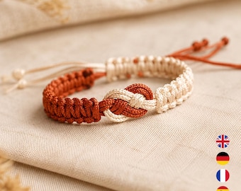 Könnte beinhalten: Ein Makramee-Armband mit einem zentralen Knotendesign und abwechselnden Abschnitten aus rostorangefarbener und cremefarbener Kordel. Das Armband verfügt über verstellbare Zugbänder mit kleinen Knoten an den Enden und eine durchgehend gewebte Struktur.